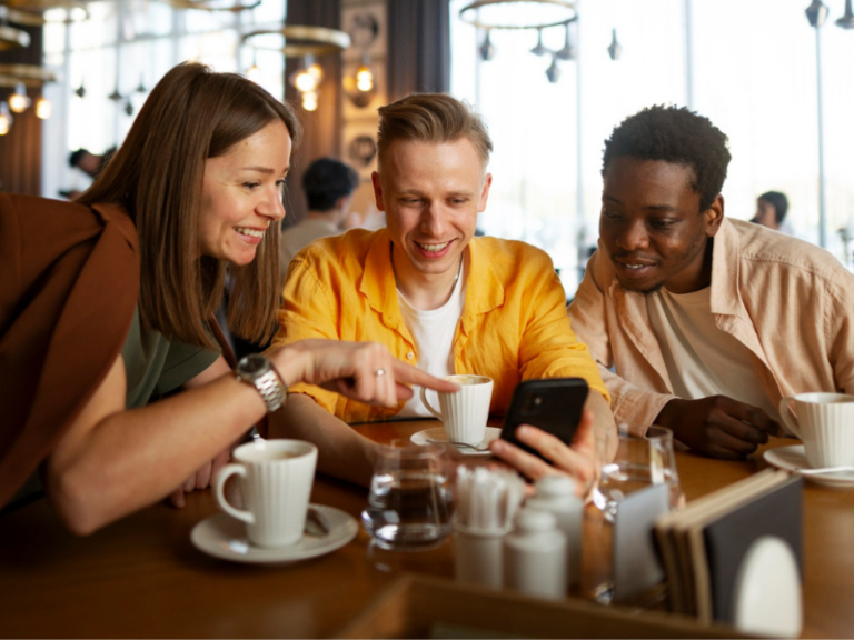 Three friends sitting together at a café table, looking at a smartphone one of them is holding, surrounded by coffee cups and glasses, in a bright, modern café setting with natural light and circular ceiling lights.